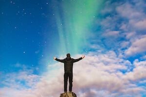 man standing on rock under green aurora borealis and white clouds